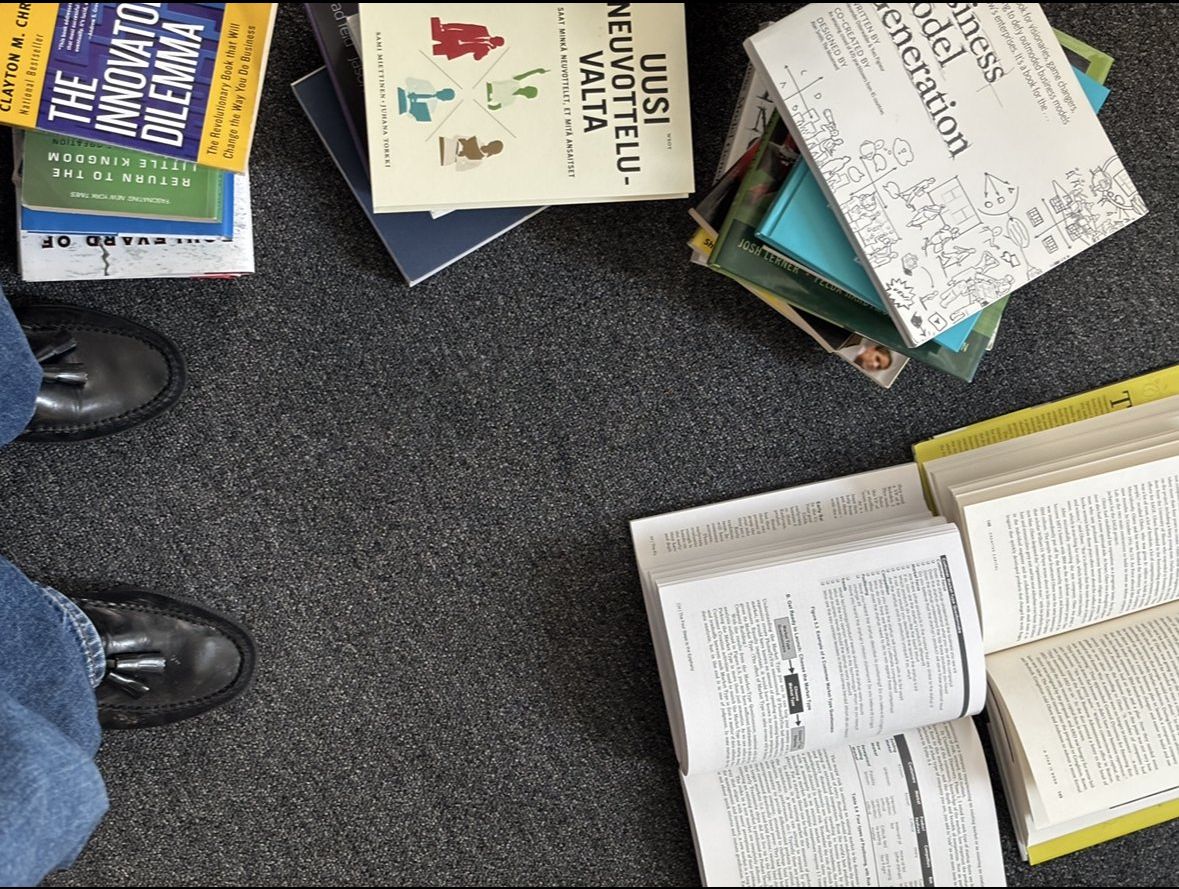 Overhead view of the archive: open books, a stack of titles, denim and a leather shoe on a dark carpet.