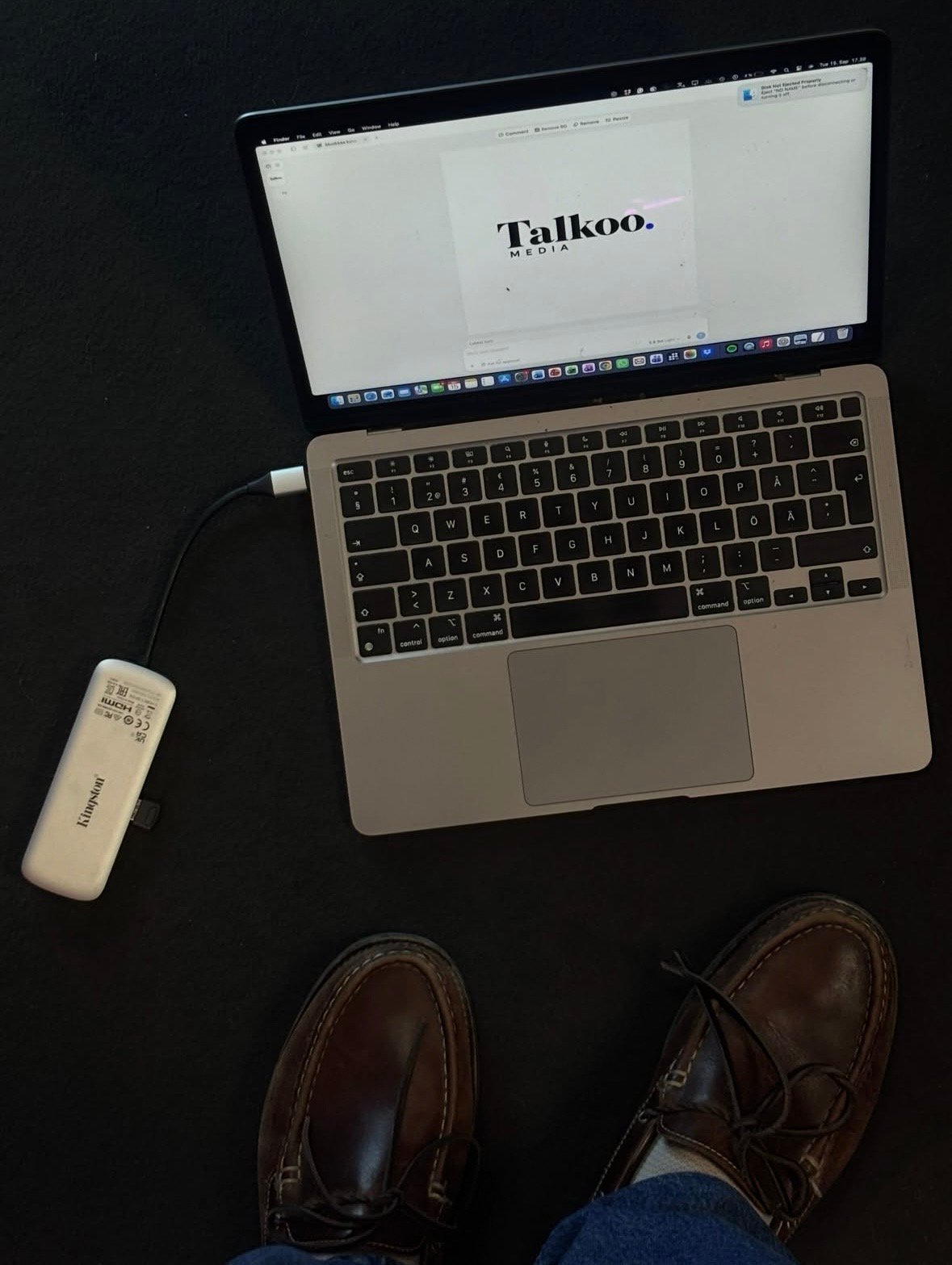 Behind the scenes: a laptop showing the Talkoo Media wordmark on a dark floor, next to a USB drive and the photographer's shoes.