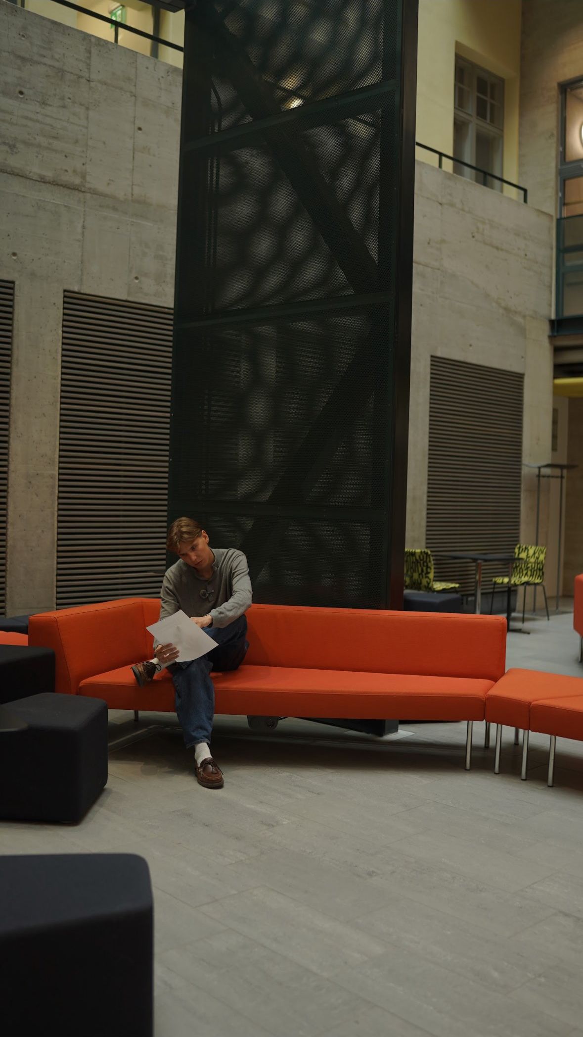 A builder reading a script on an orange sofa in a concrete lobby.