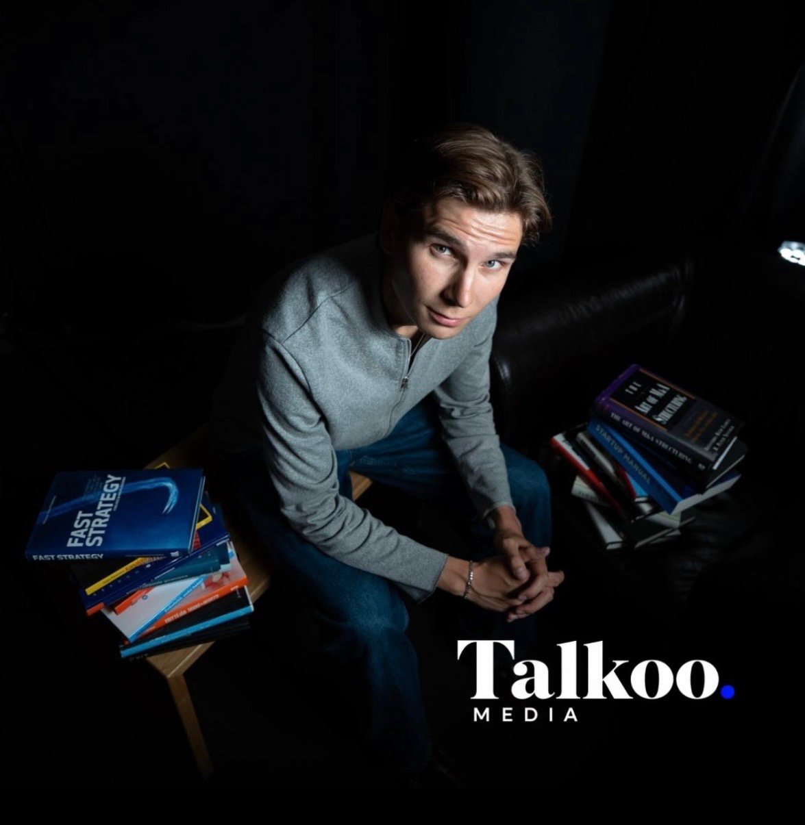 A builder sitting among stacks of books in a dark room, Talkoo Media wordmark in the corner.